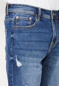 Modre jeans hlače iz-denima z obrabljenimi detajli, s ravnim krojem, rumenim šivanjem ter vidnimi sprednjimi žepi in kovinskimi dodatki.
