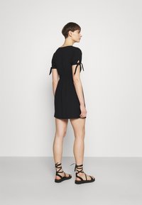 Vestido curto preto com mangas bufantes e detalhes em laço, apresentando uma cintura justa e saia rodada. Combinado com sandálias pretas de tiras.