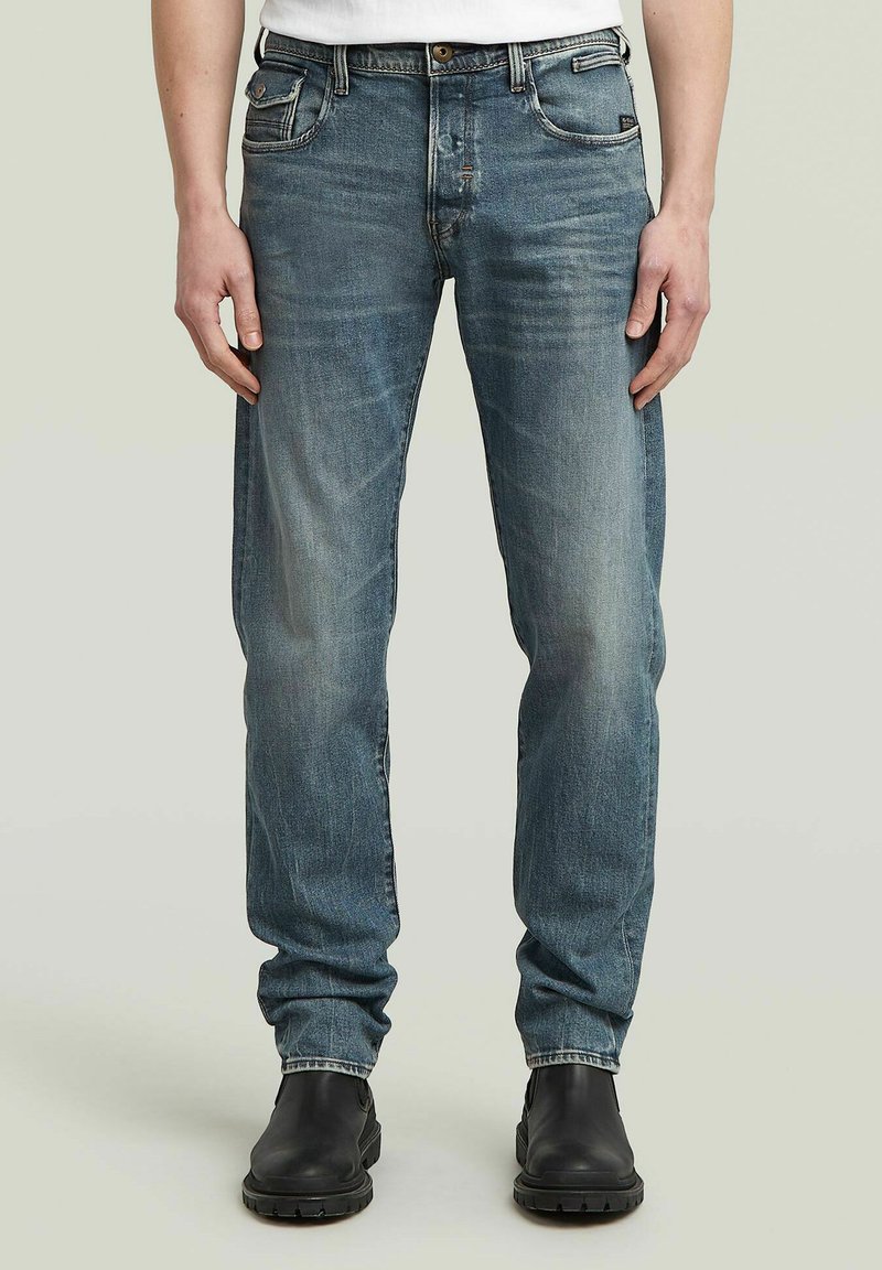 Homem vestido com jeans azuis desbotados e botas de couro pretas, em pé com as mãos relaxadas ao lado do corpo, contra um fundo neutro.