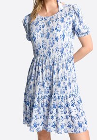 Vestido azul con estampado floral que presenta un diseño en capas, mangas cortas abullonadas y textura acanalada. La tela tiene un fondo blanco con patrones azules.