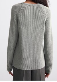 Femme aux cheveux bouclés portant un pull en maille côtelée gris clair et un pantalon à rayures grises foncées, vue de dos.