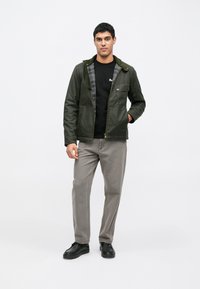 Jachetă verde măsliniu cu fermoar, material texturat, căptușeală în carouri și elemente metalice aurii. Asortată cu pantaloni gri și pantofi negri.