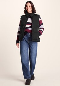 Smiling woman wearing a black puffer vest over a multicolored striped sweater, high-waisted blue jeans, and black ankle boots.