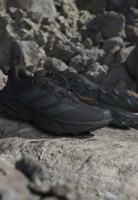Paire de chaussures de trail running athlétiques noires sur un terrain rocheux, avec semelles texturées et design à lacets.