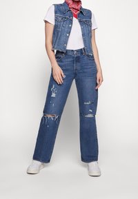 Chaleco de mezclilla con bordes desgastados, camiseta blanca y jeans de pierna ancha en azul claro con desgastes y roturas. Zapatillas blancas completan el atuendo.