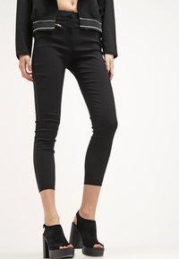 Kvinna i svarta avkortade skinny jeans med dragkedjefickor och svarta plattformssandaletter med öppen tå, stående mot en vit bakgrund.