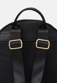Sac à dos en tissu noir avec une texture lisse, doté de quincaillerie couleur or et de bretelles noires ajustables fixées par des boucles carrées.