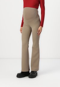 Zwangere flared broek in beige katoenmix met een hoge, rekbare tailleband. Gecombineerd met een aansluitend rood T-shirt met lange mouwen en zwarte stevige laarzen.