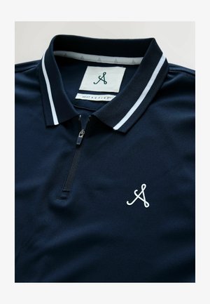 Granatowa koszulka polo z połowicznym zamkiem błyskawicznym, kołnierzykiem w białe paski i białym logo. Wykonana z gładkiego, strukturalnego materiału. Rozszycia widoczne wewnątrz.