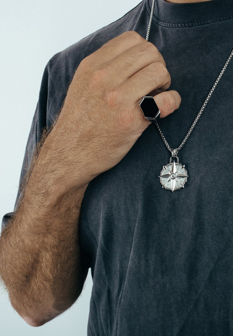 Collier en argent avec pendentif en forme de boussole, présentant des bords tranchants et une pierre centrale. Main portant une bague hexagonale noire sur une chemise grise.