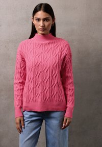 Růžový pletený svetr s vysokým límcem, který má texturovaný diamantový vzor, ribbed manžety a spodní lem, spárovaný s světle modrými džíny.