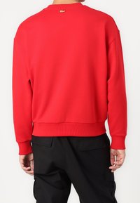 Sudadera roja con cuello redondo, puños y dobladillo acanalados. Presenta un pequeño logo dorado cerca del cuello. Textura de tela lisa. Pantalones negros visibles.