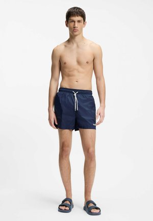 Młody mężczyzna stoi boso, ubrany w granatowe spodenki kąpielowe ze białym sznurkiem oraz granatowe klapki, na prostym białym tle.