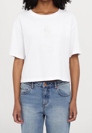 Kvinde iført en løs hvid cropped T-shirt med subtilt præget logo og lyseblå jeans med høj talje mod en ensfarvet baggrund.