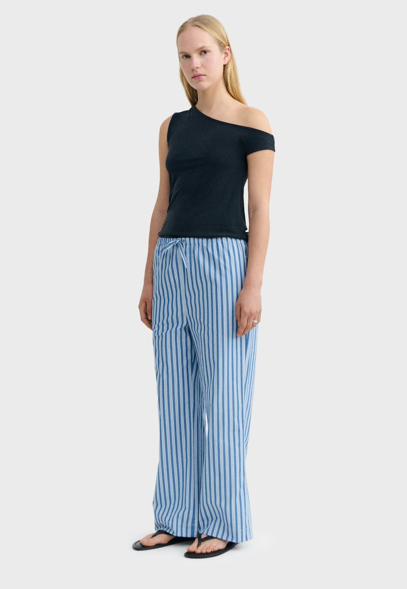 Femme portant un haut noir à une épaule avec un pantalon large à rayures verticales bleues et blanches et des tongs noires, debout devant un fond uni.