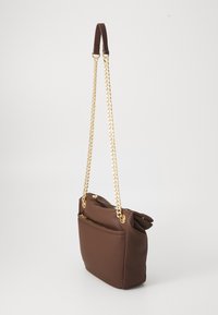 Braune Schultertasche mit goldenen Kettenriemen und einer vorderen Reißverschlusstasche, die an einem schlichten weißen Hintergrund hängt.
