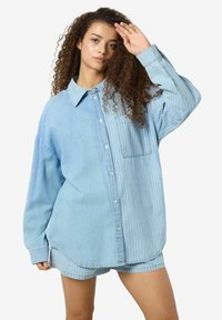 Chemise en denim bleu clair oversize avec un motif de rayures verticales d'un côté, fermeture à boutons et une poche poitrine.