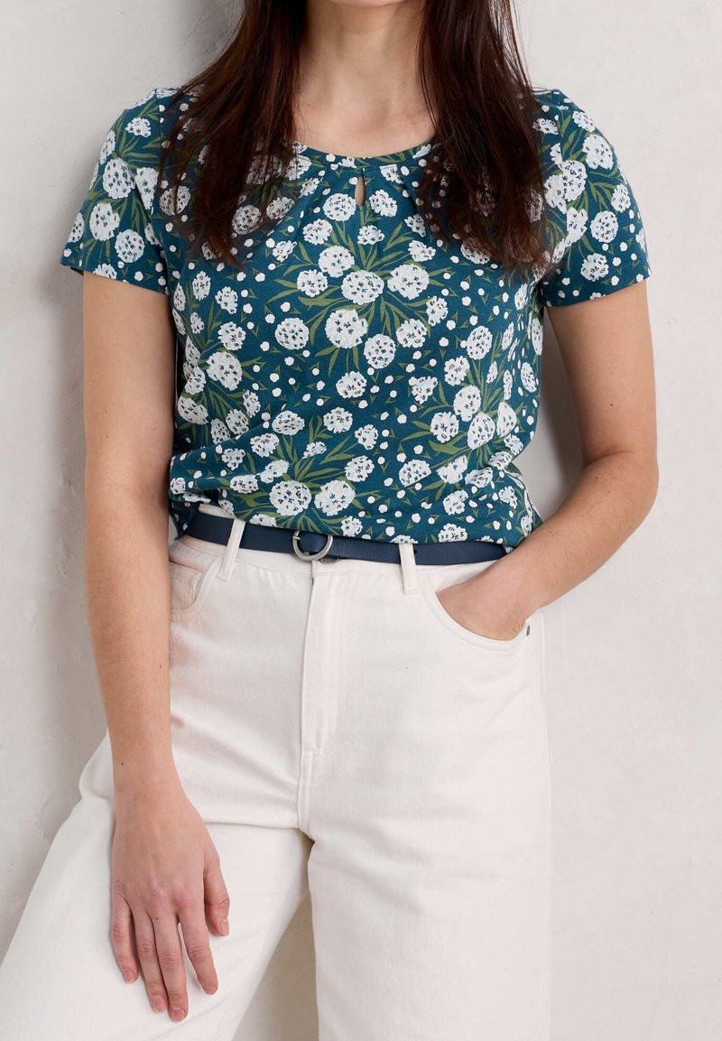 Krátký rukáv, tyrkysový květinový top s bílými květy a zelenými listy, doplněný o vysoké béžové kalhoty a námořnický pásek.