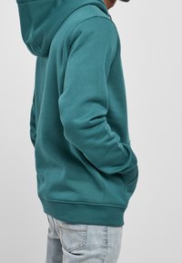 Sudadera con capucha color azul verdoso, con bolsillo canguro, puños y bajo acanalados. Textura de tela suave, capucha adjunta, combinada con vaqueros azul claro.