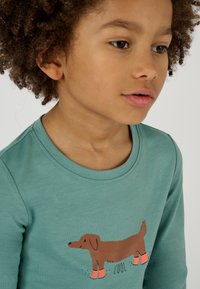 Maglietta a maniche lunghe color teal con scollatura rotonda, caratterizzata da una grafica di un cane marrone che indossa stivali rosa e la parola "COOL" sotto.