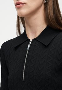 Γυναίκα που φοράει μαύρο υφασμάτινο τοπ με ασημένιο φερμουάρ και γιακά, εμφανίζοντας το κάτω μέρος του προσώπου, το λαιμό και καστανά μαλλιά πάνω σε λευκό φόντο.