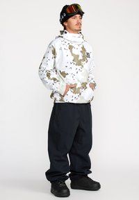 Sudadera blanca con patrón de camuflaje, con un alto cuello y bolsillos, combinada con pantalones impermeables negros y botas de nieve negras.