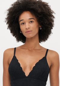 Fekete csipkés bralette mély V-nyakkal, állítható pántokkal, virágmintával és fodros szélekkel. Sima textúrájú, testhez simuló kialakítással.