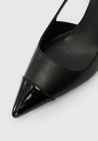 Musta teräväkärkinen kenkä, jossa on sileä nahka, kiiltävä patenttikoriste kärjessä ja avoimet leikkauskuviot sivuilla.