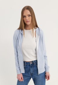 Sudadera de cremallera azul claro con capucha ajustable, puños acanalados y dos bolsillos frontales, combinada con jeans de talle alto y una camiseta blanca.