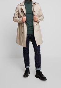 Beige Trenchcoat mit doppelreihigen Knöpfen, langen Ärmeln und Schulterklappen, getragen über einem dunklen grünen Rollkragenpulli, kombiniert mit dunklen Jeans und schwarzen Schuhen.