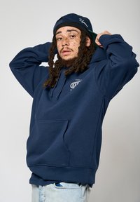 Sweatshirt à capuche bleu marine avec une poche avant et un logo. Présente une coupe décontractée et des poignets côtelés. Porté avec une casquette noire.