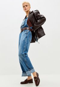 Žena u smeđoj kožnoj jakni preko traper kombinezona s širokim smeđim remenom i tamno smeđim cipelama, pozira ispred bijele pozadine.