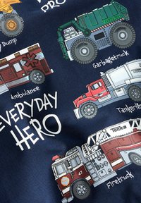 Tessuto navy con illustrazioni colorate di vari camion: camion dei pompieri, ambulanza, camion della spazzatura e autocisterna, etichettati con i nomi.