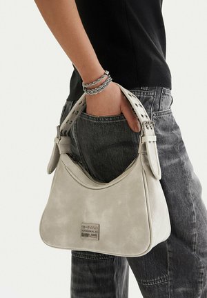 Mano sosteniendo un bolso de hombro de color beige claro de G-Star Originals, llevando pulseras trenzadas plateadas, camiseta negra y vaqueros negros descoloridos.