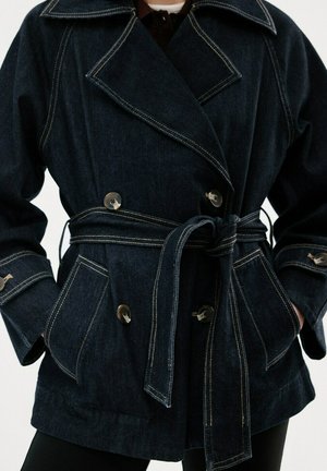 Ciemnogranatowa kurtka dżinsowa z paskiem, zapinana na podwójne guziki, z szerokimi klapami i przednimi kieszeniami na klapę, noszona na czarnych spodniach.