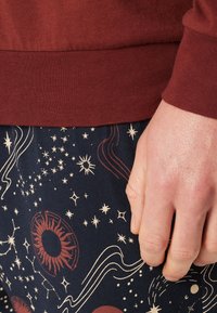 Huppari, jossa on tummanpunainen helma ja ribbikudos, yhdistettynä laivastonsinisiin housuihin, joissa on kultaiset taivaankappalepatternit, mukaan lukien tähtiä ja aurinkomotifeja.