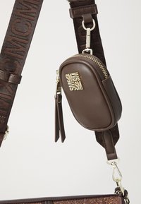 Sac en cuir marron avec une fermeture éclair dorée, pochette plus petite attachée et bandoulière marron avec motif de logo. Texture lisse et accents métalliques.