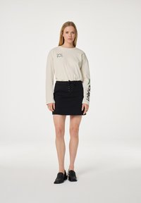 Langarmige crèmekleurige shirt met groene graphics, gecombineerd met een zwarte spijkerrok met vetersluiting aan de voorkant. Zwarte instappers maken de outfit compleet.