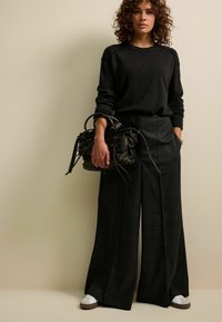 Schwarzer Strickpullover mit gerippten Bündchen, kombiniert mit weit geschnittenen karierten Hosen in Dunkelgrau. Schwarze Handtasche mit strukturierten Details und weißen Sneakers.