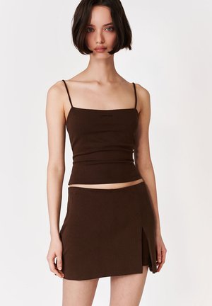 Młoda kobieta z krótkimi ciemnymi włosami, ubrana w ciemnobrązowy top na cienkich ramiączkach oraz dopasowaną mini spódnicę kopertową, stoi na jednolitym tle.