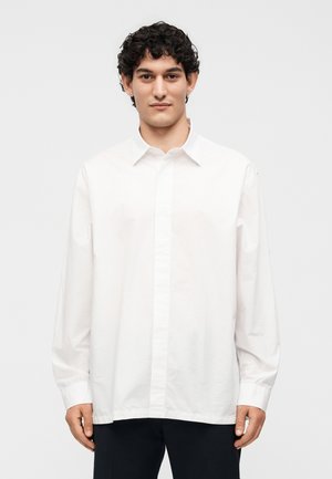 Άνδρας με σγουρά σκούρα μαλλιά που φοράει μια άνετη λευκή μπλούζα με μακριά μανίκια και κουμπιά και μαύρο παντελόνι, στέκεται σε λευκό φόντο.