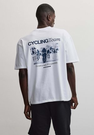 Homme en t-shirt blanc avec le texte "CYCLING Team" et un dessin d'un groupe de cyclistes ; le slogan indique : "It's not a race. It's a journey."
