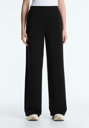 Frau trägt hochgeschnittene, weit geschnittene schwarze Hose kombiniert mit weißen Turnschuhen und einem in die Hose gesteckten weißen Oberteil.