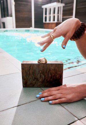 Main avec vernis à ongles bleu et bagues en or qui dépasse d'une pochette en marbre au bord de la piscine, reposant sur un sol carrelé.