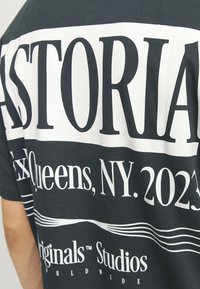 Tamsiai pilka marškinėliai su ryškiu baltu tekstu. Tekstas apima "ASTORIA", "Queens, NY. 2023" ir "Originals Studios" su dryžuotu dizainu.