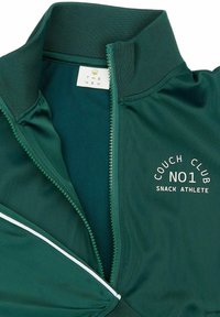 Giacca verde con zip e colletto alto. Presenta una striscia di accento bianca e la scritta stampata "COUCH CLUB NO1 SNACK ATHLETE" sul petto.