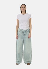 Woman wearing a fitted white short-sleeve shirt and light blue wide-leg jeans with a unique overlapping waistband, standing against white background.