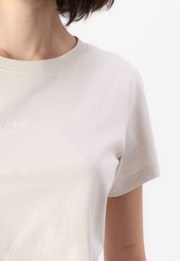Beige cotton t-shirt with a crew neckline and short sleeves. Features embossed logo detailing across the chest.