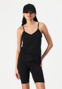 Canottiera nera leggera con spalline sottili, abbinata a pantaloni ciclismo neri. Il tessuto è liscio, creando una silhouette aderente.