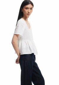 Blouse blanche texturée avec un col en V et des manches courtes, au design peplum flottant, associée à un jean en denim sombre à jambes larges.